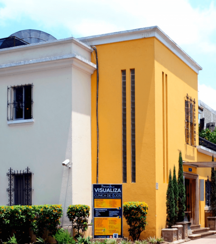 Fotografía de nuestra clínica privada en la zona 9 visualiza casa de dos niveles con el frente color amarillo y de fondo lado izquierdo la pared color blanco hueso rodeada por plantas